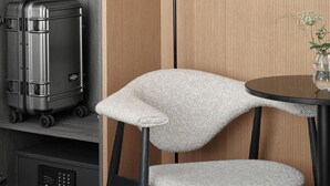 Ein kleines Zimmer mit einem grauen Koffer, einem Stuhl, einem kleinen Tisch mit einer Vase und einer Stehlampe.