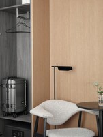 Habitación pequeña con una maleta gris, una silla, una mesita con un florero y una lámpara de pie.