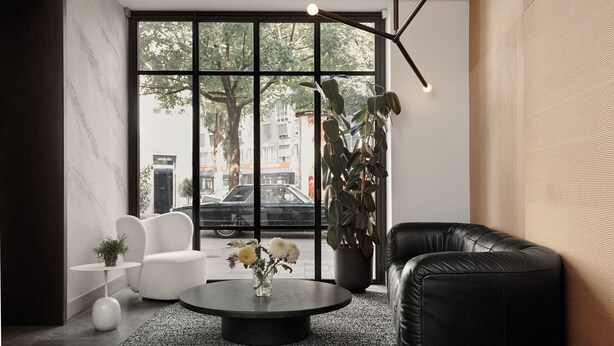 Ein modernes Interieur mit einer schwarzen Ledersofa, einem weißen Sessel und einem runden Couchtisch mit Blumen. Große Fenster bieten Blick auf die Straße.
