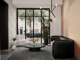 Un interior moderno con un sofá de cuero negro, un sillón blanco y una mesa de centro redonda con un jarrón de flores. Amplias ventanas ofrecen vistas a la calle.