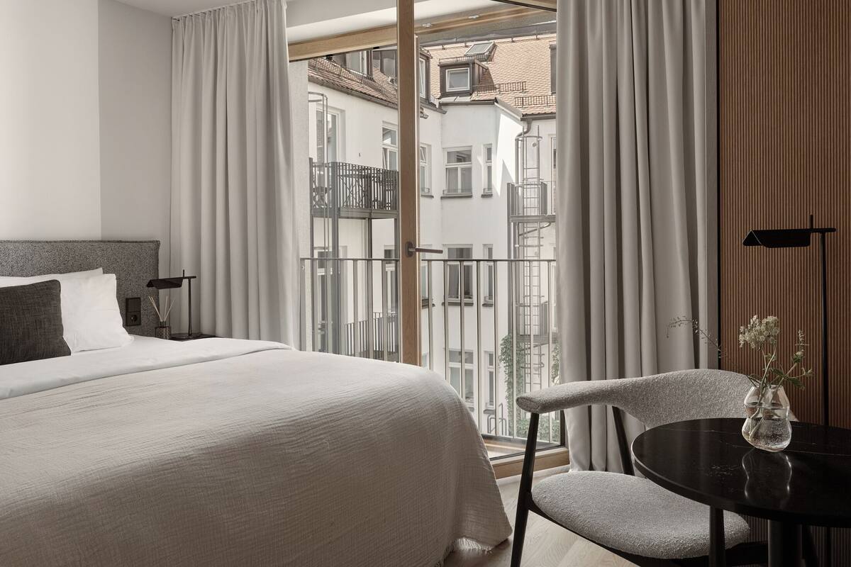 A hotel room with a bed, a chair, a table, and a view of the city through large windows.