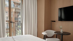 Ein Hotelzimmer mit einem großen Bett, einem Sessel, einem kleinen Tisch, einem Fernseher und Blick auf die Stadt durch ein Fenster.