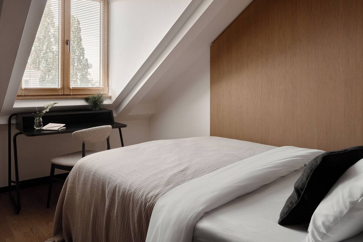 A bedroom with a sloped ceiling, a desk with a chair, a bed with white bedding, and a window with blinds.