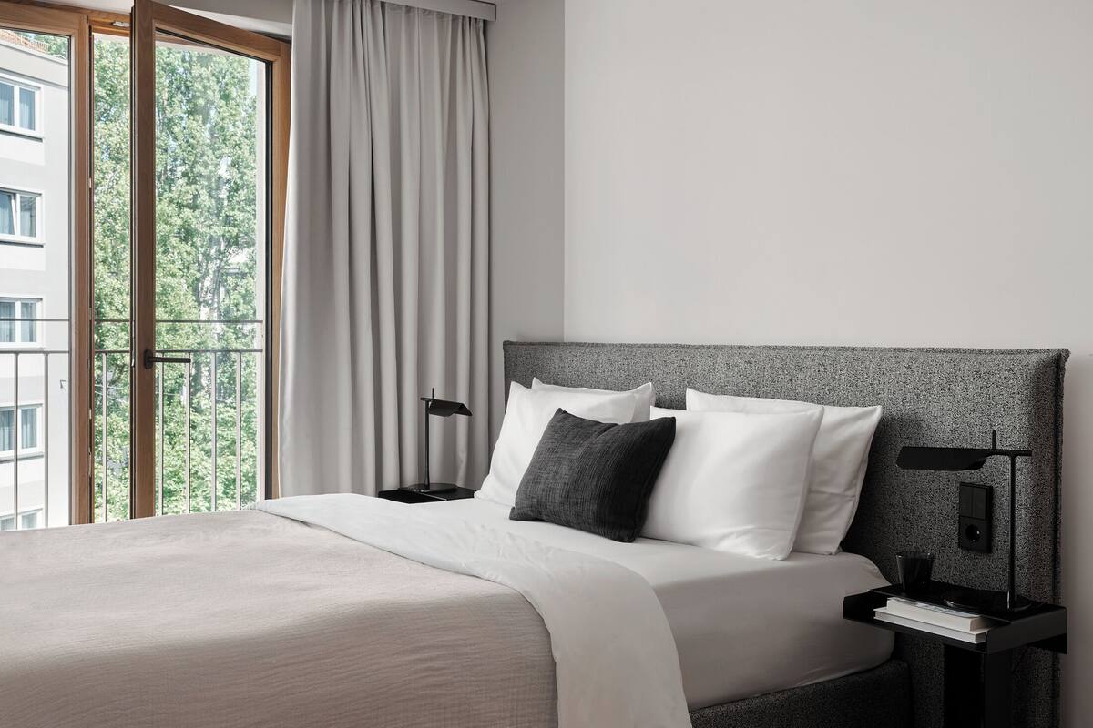 A modern hotel room with a large bed, a nightstand, a reading lamp, and a view of trees through the window.