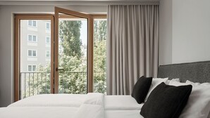 Ein Hotelzimmer mit einem großen Bett, Blick auf ein Gebäude und Bäume durch ein Fenster sowie einem Balkon.