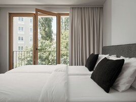 Habitación de hotel con una cama grande, vistas a un edificio y árboles a través de una ventana, y un balcón.