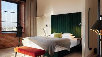 Habitación de hotel con un ventanal grande, un maniquí, una cama con cabecera verde, un banco y un escritorio con teléfono.