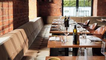 A restaurant with a long wooden table set for dining, a cushioned bench along the wall, and a brick wall.