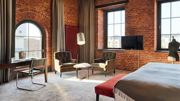 A hotel room with brick walls, a large window, a desk, two armchairs, a red ottoman, and a bed.