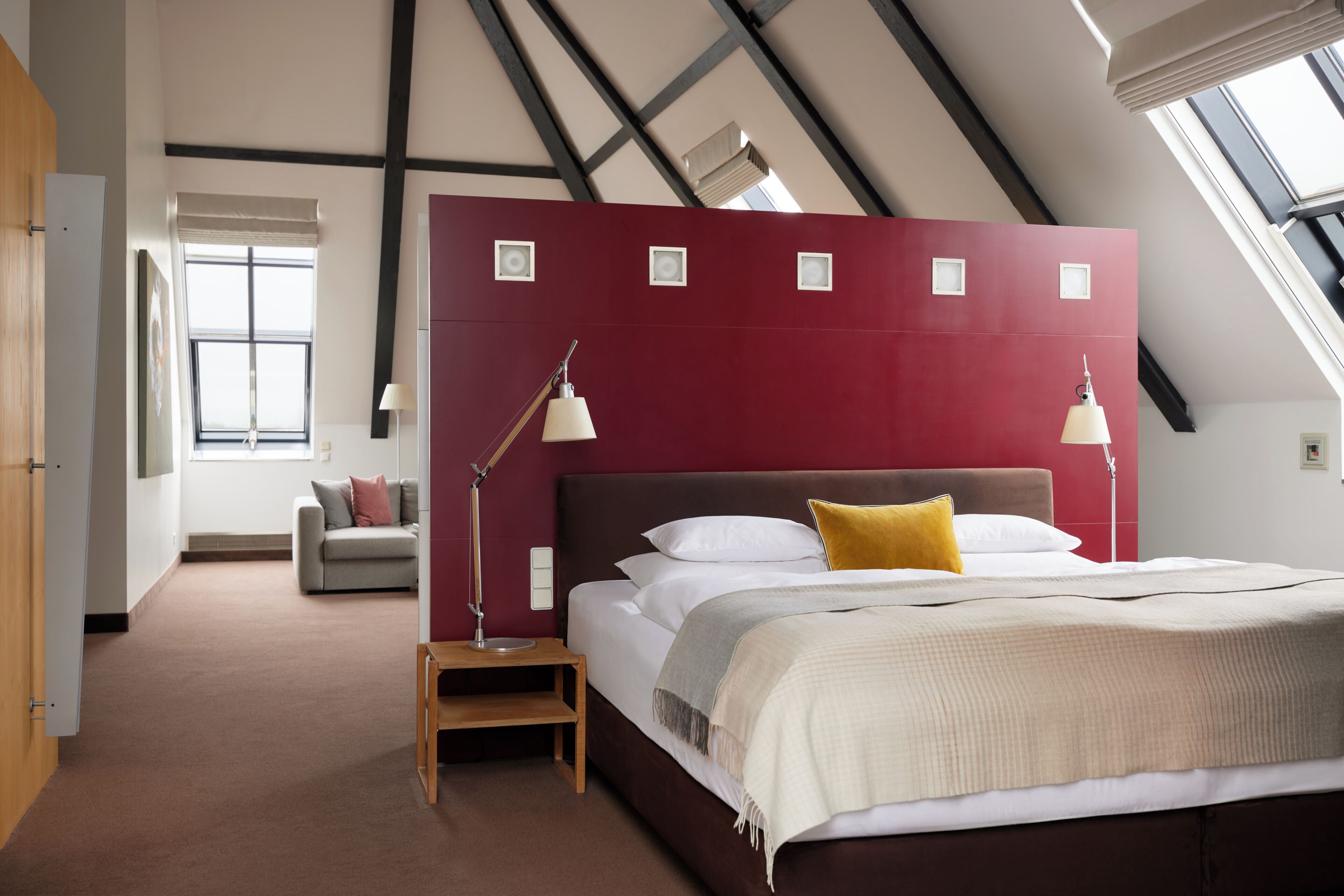 Ein Schlafzimmer mit schrägem Dach, einem Bett mit weißen Leinen, einer roten Akzentwand und einem Fenster, das natürliches Licht hereinlässt.