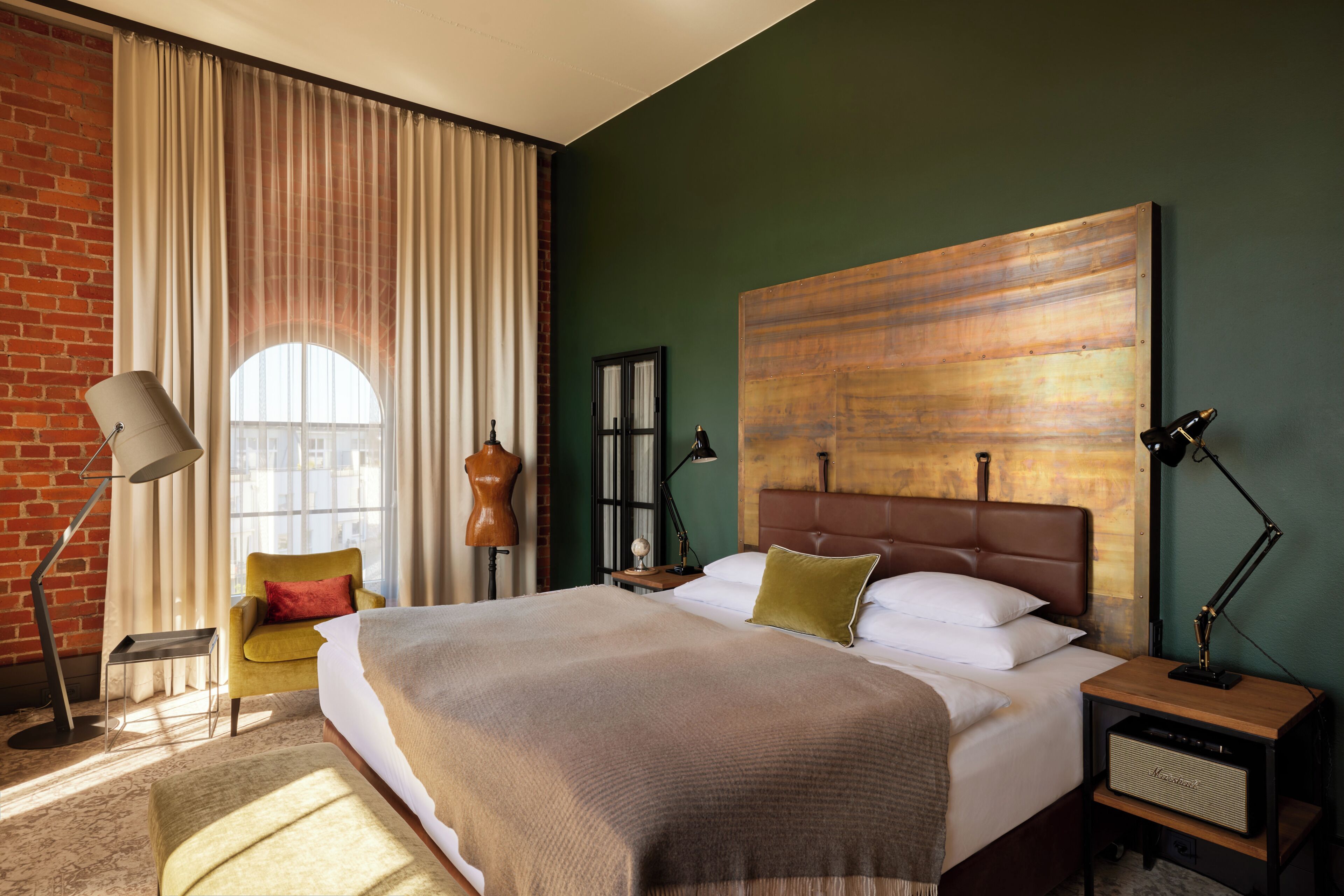 Ein Hotelzimmer mit einem großen Bett, einem Schreibtisch mit Lampe, einem Sessel und einem großen Fenster mit Vorhängen.