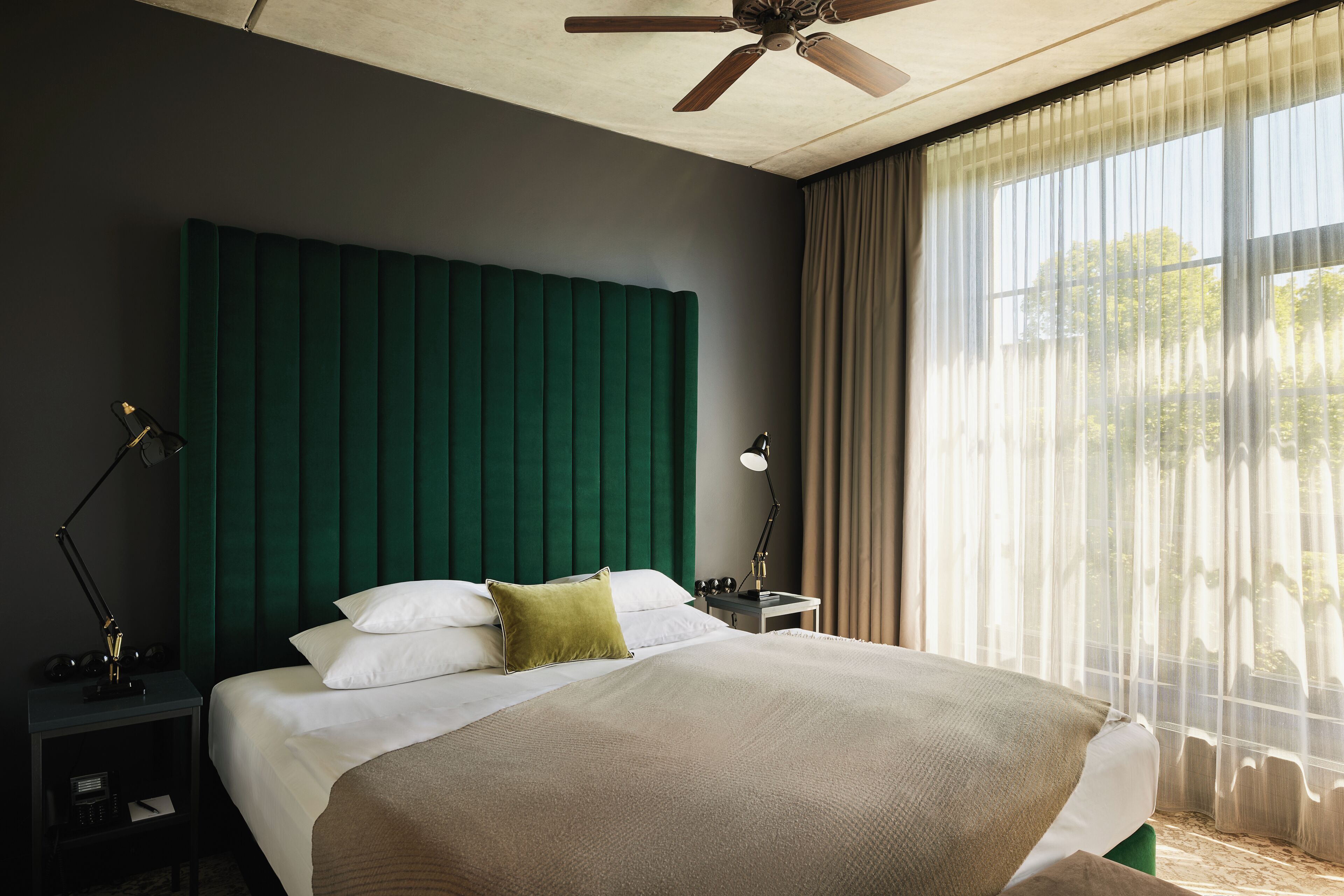 Een hotelkamer met een groot bed, een groen hoofdeinde, een nachtkastje met een lamp en een raam met dunne gordijnen.