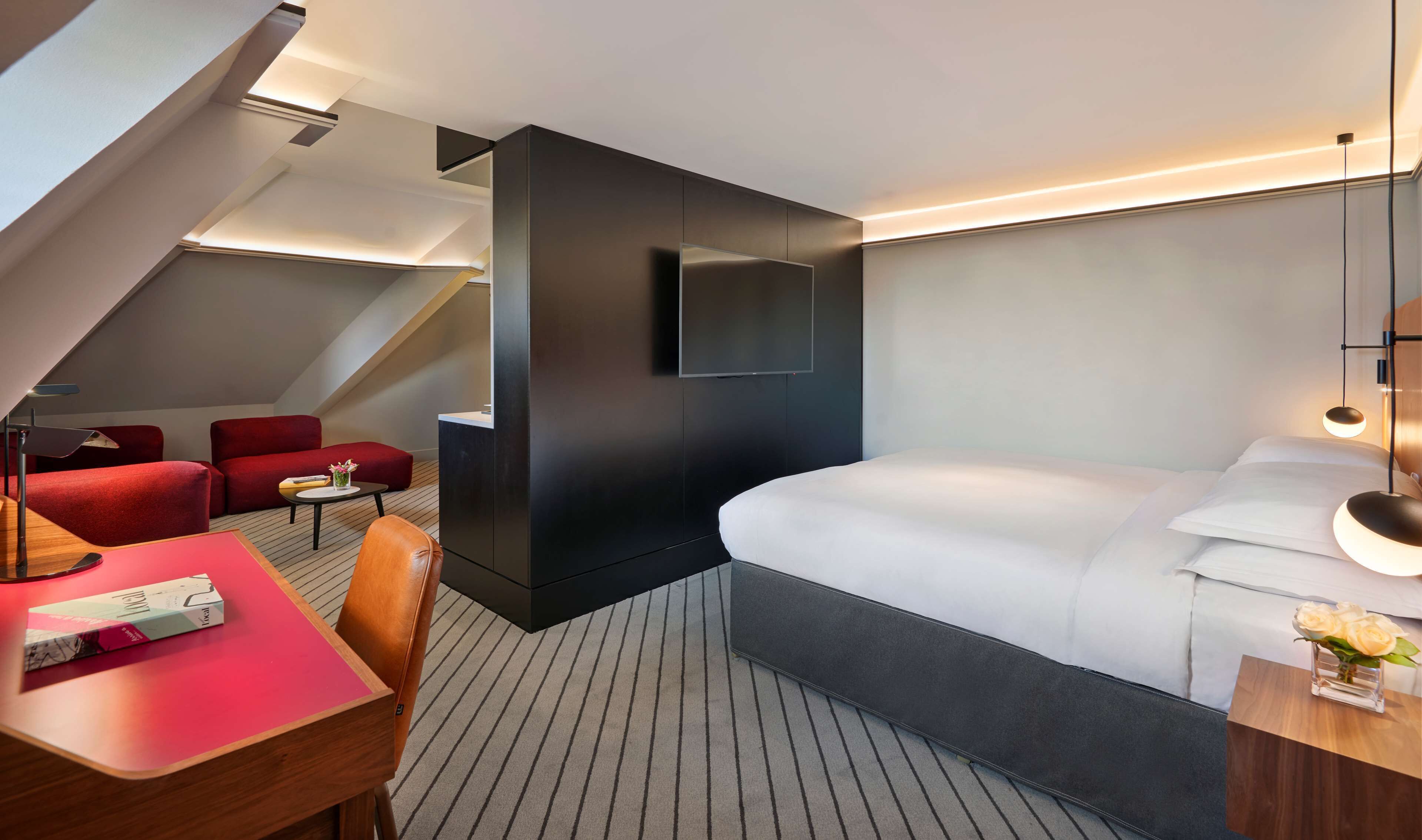 A modern hotel room with a large bed, a desk with a chair, a small table, and two red sofas.
