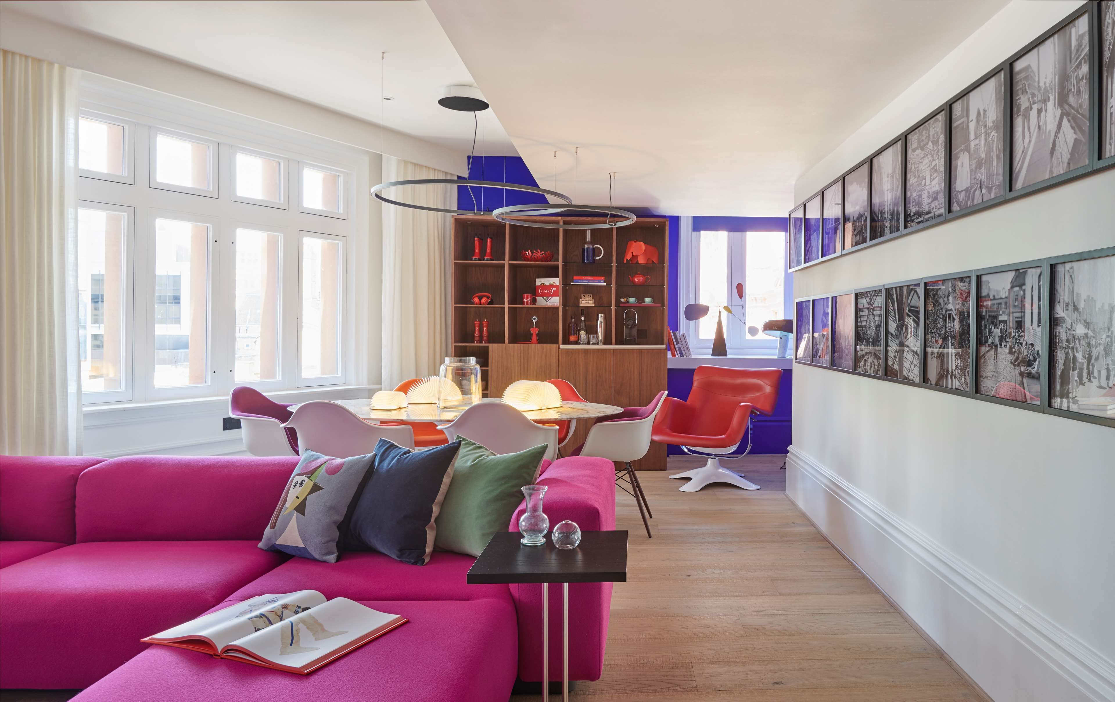 Un salon moderne avec un canapé rose vif, un coin repas doté d’une table et de chaises en bois, et une bibliothèque remplie de divers objets.