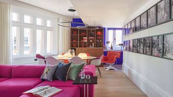 Un salon moderne avec un canapé rose vif, un coin repas doté d’une table et de chaises en bois, et une bibliothèque remplie de divers objets.