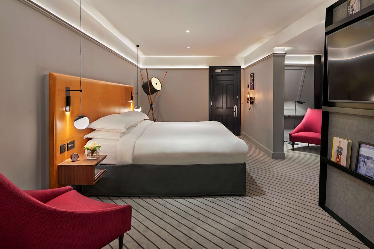 Een moderne hotelkamer met een groot bed, een houten hoofdbord, een rode fauteuil, een kleine tafel met een klok en een vaas met bloemen, en een televisie op een dressoir.