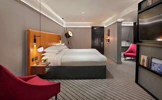 Una habitación de hotel moderna con una cama grande, cabecera de madera, un sillón rojo, una mesita con un reloj y un jarrón de flores, y un televisor sobre un mueble.