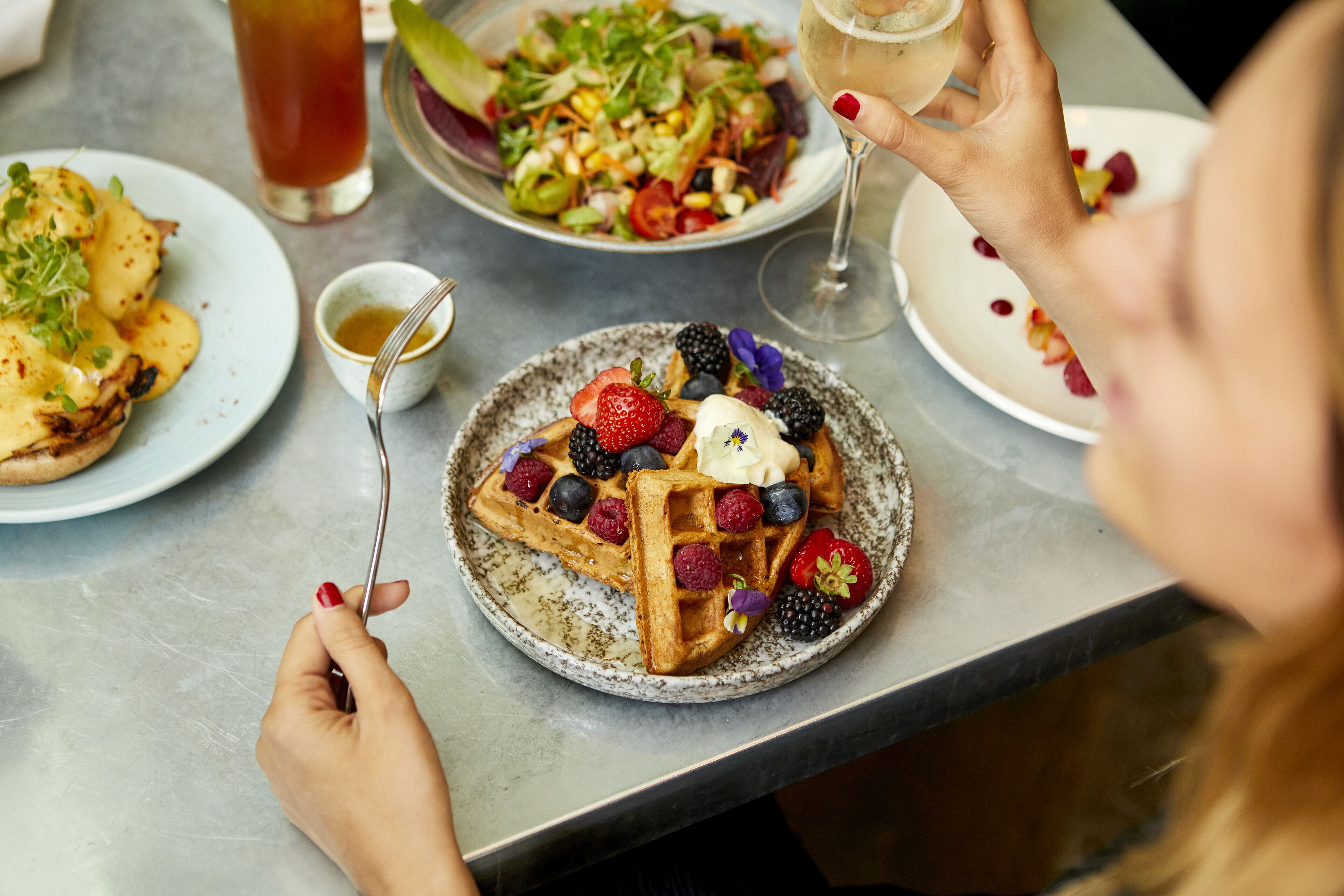 Une table à manger avec des assiettes remplies de nourriture, notamment des gaufres garnies de fruits rouges et de crème fouettée, une salade et un verre de thé glacé.