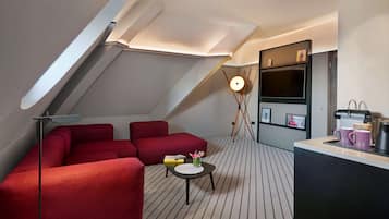 A modern loft bedroom with a red sectional sofa, a small round table with a vase, a floor lamp, a TV, and a kitchenette.
