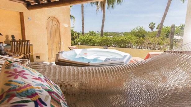 Een hangstoel met een hottub op de achtergrond, een houten deur en uitzicht op palmbomen en groen.
