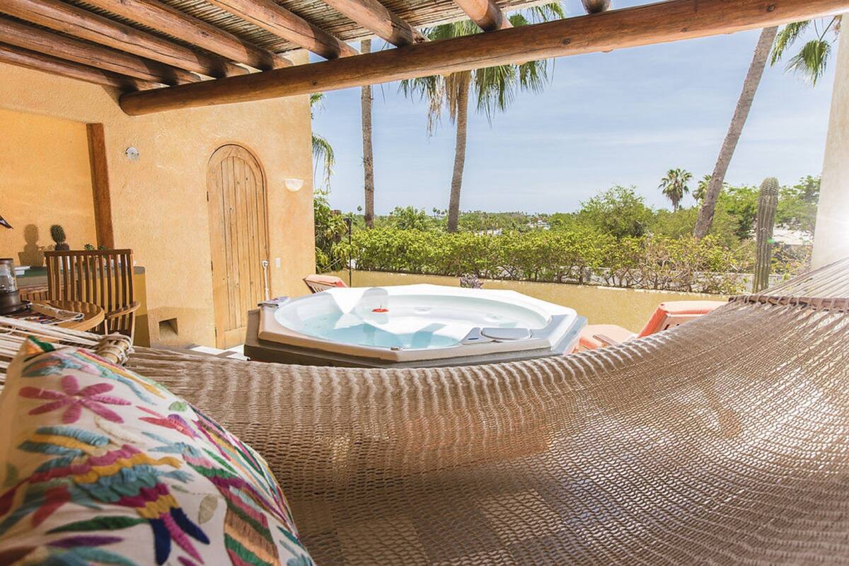 Een hangstoel met een hottub op de achtergrond, een houten deur en uitzicht op palmbomen en groen.