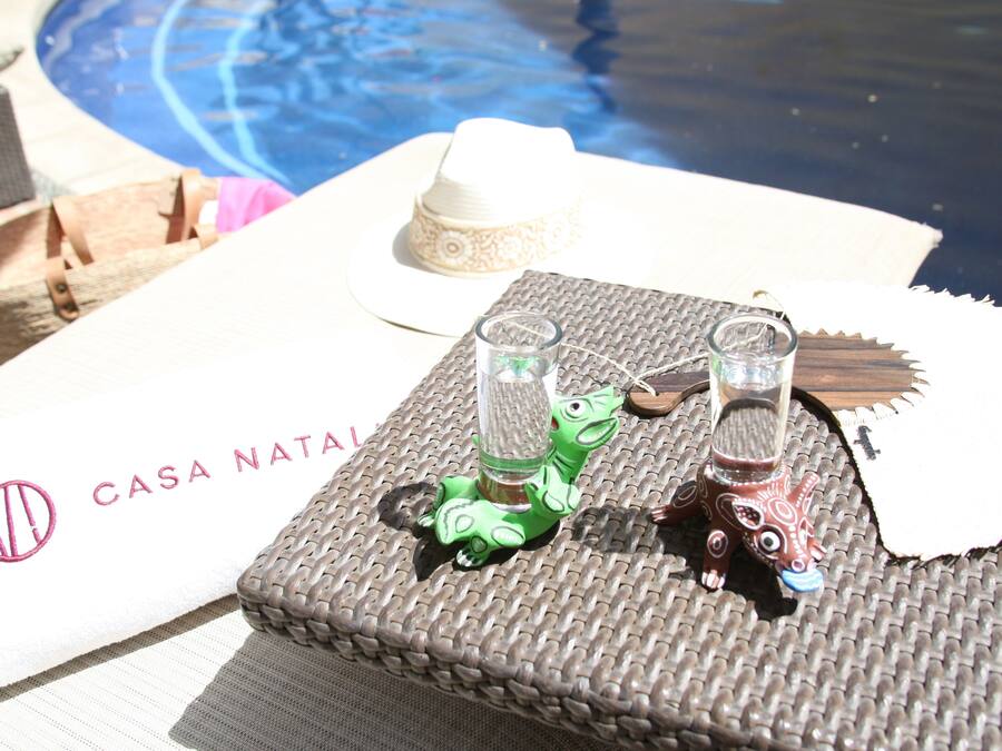 A poolside setting with a woven mat, two small glass bottles, a hat, and a bag on a white cloth with "CASA NATAL" printed on it.