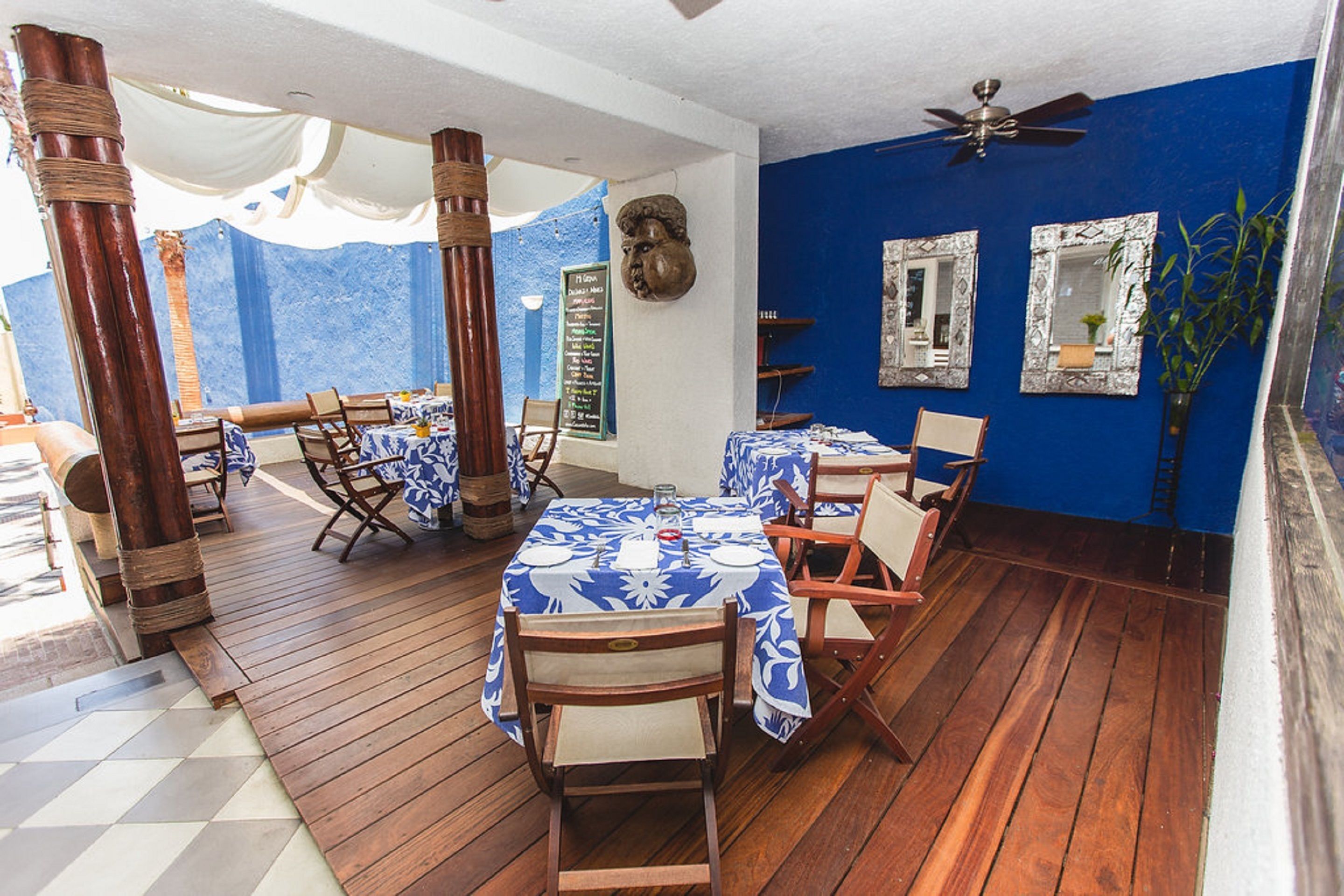A restaurant with wooden flooring, blue walls, and white ceiling. Tables are set with blue tablecloths and white chairs.