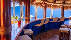 Een bar met een gebogen houten toonbank, rieten stoelen en uitzicht op palmbomen.