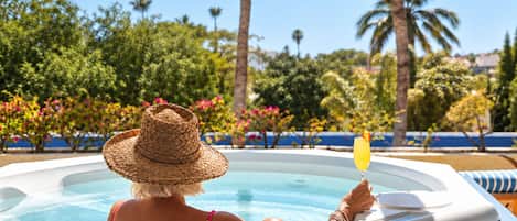 Une personne se détend dans un bain à remous, un verre à la main, entourée d’une végétation luxuriante et de palmiers.