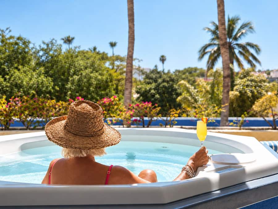 A person relaxing in a hot tub with a drink, surrounded by lush greenery and palm trees.