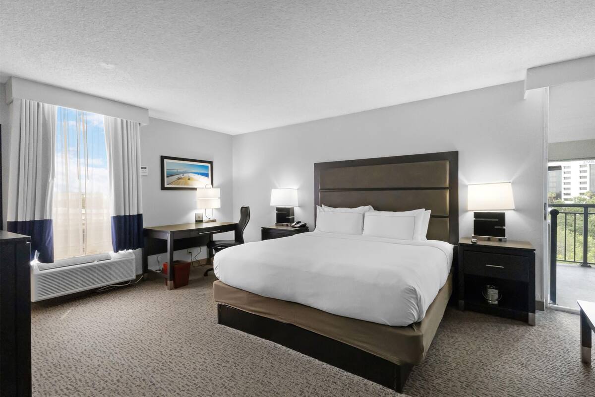 Une chambre d’hôtel avec un grand lit, un bureau, deux tables de chevet avec des lampes, une chaise, une fenêtre avec des rideaux et un tableau au mur.