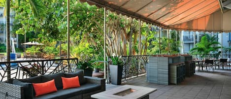 Un espacio al aire libre con mobiliario de mimbre, una mesa de centro y una pérgola que proporciona sombra.
