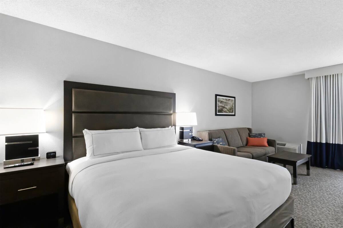 Une chambre d’hôtel avec un grand lit, un bureau avec un ordinateur, une chaise, un canapé et une fenêtre avec des rideaux.