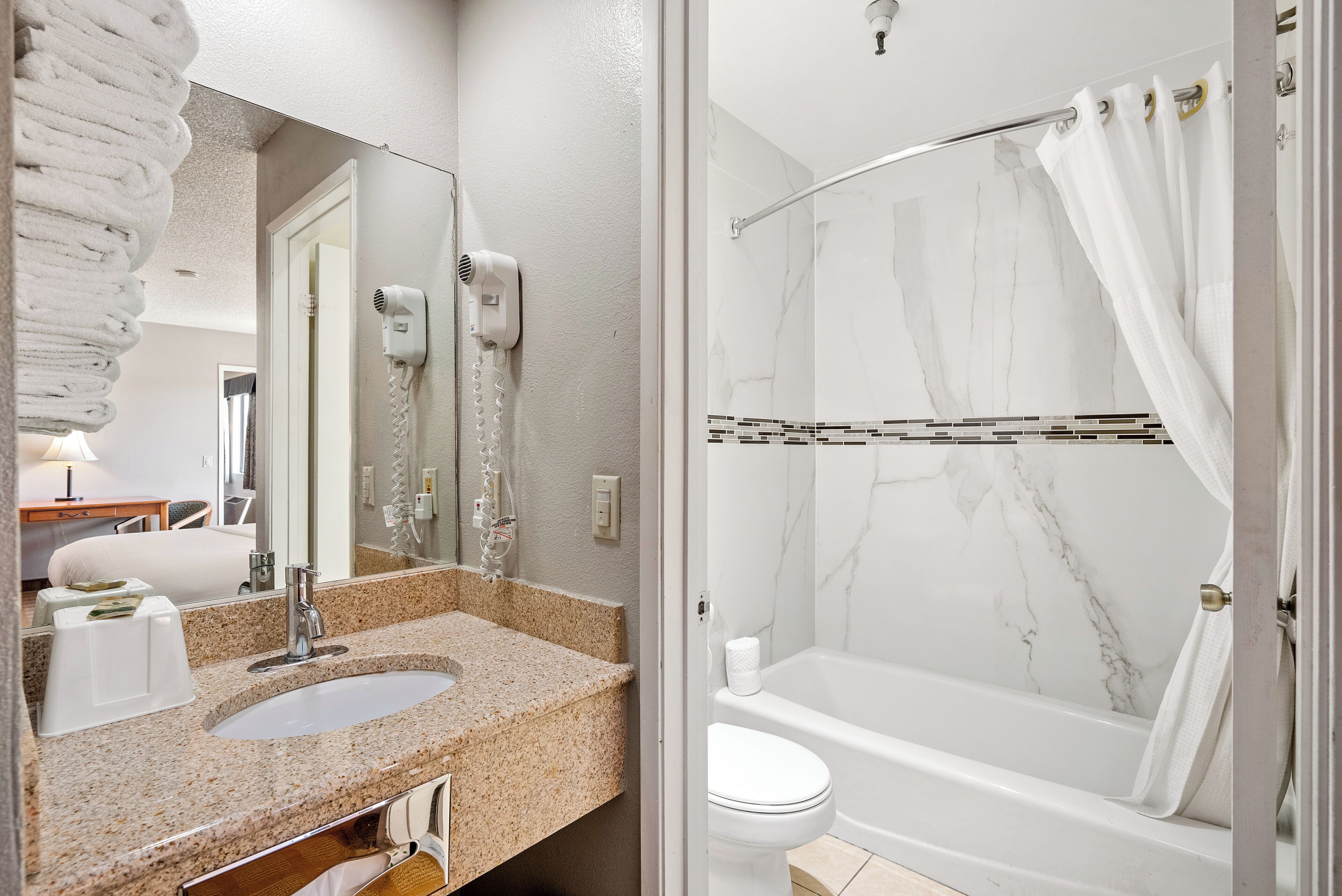 A bathroom with a granite countertop sink, a mirror, a hairdryer, a toilet, and a bathtub with a shower curtain.