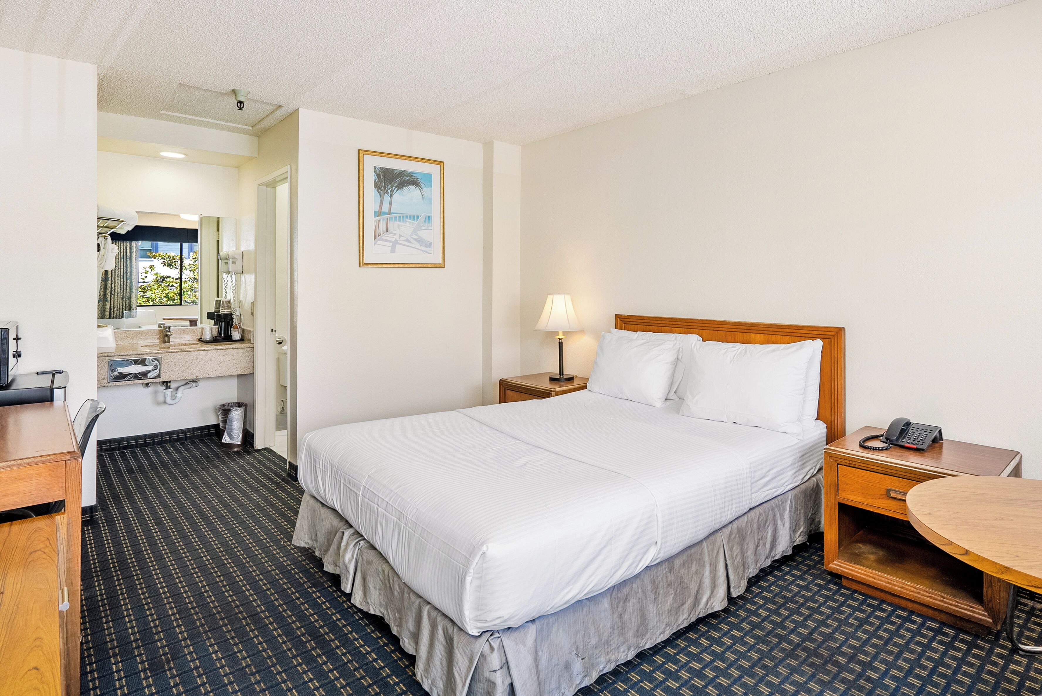 A hotel room with a large bed, a desk with a chair, a small table, a nightstand with a lamp, and a bathroom visible through an open door.