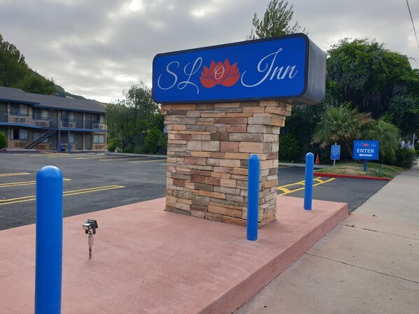 Un pilier de pierre surmonté d’une enseigne indiquant l’emplacement du SL Inn, un motel à la façade bleue, et un parking délimité par des lignes jaunes.