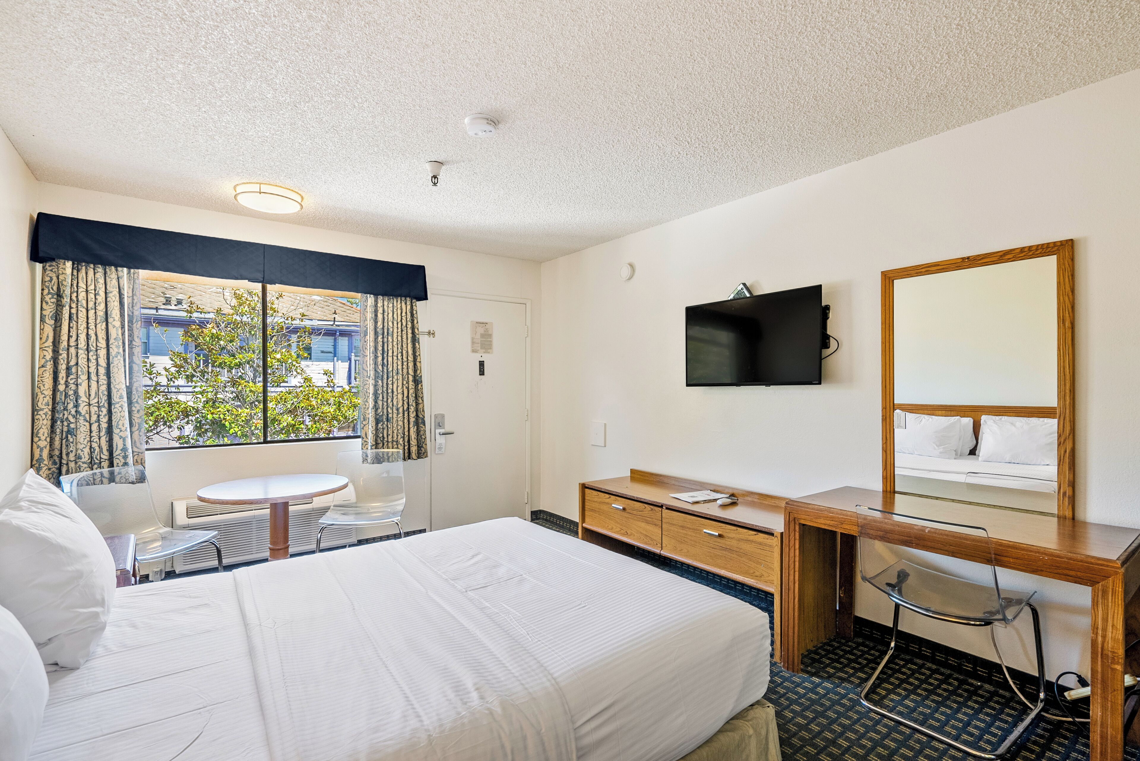 A hotel room with a bed, a desk, a chair, a round table, a mirror, and a window with curtains.
