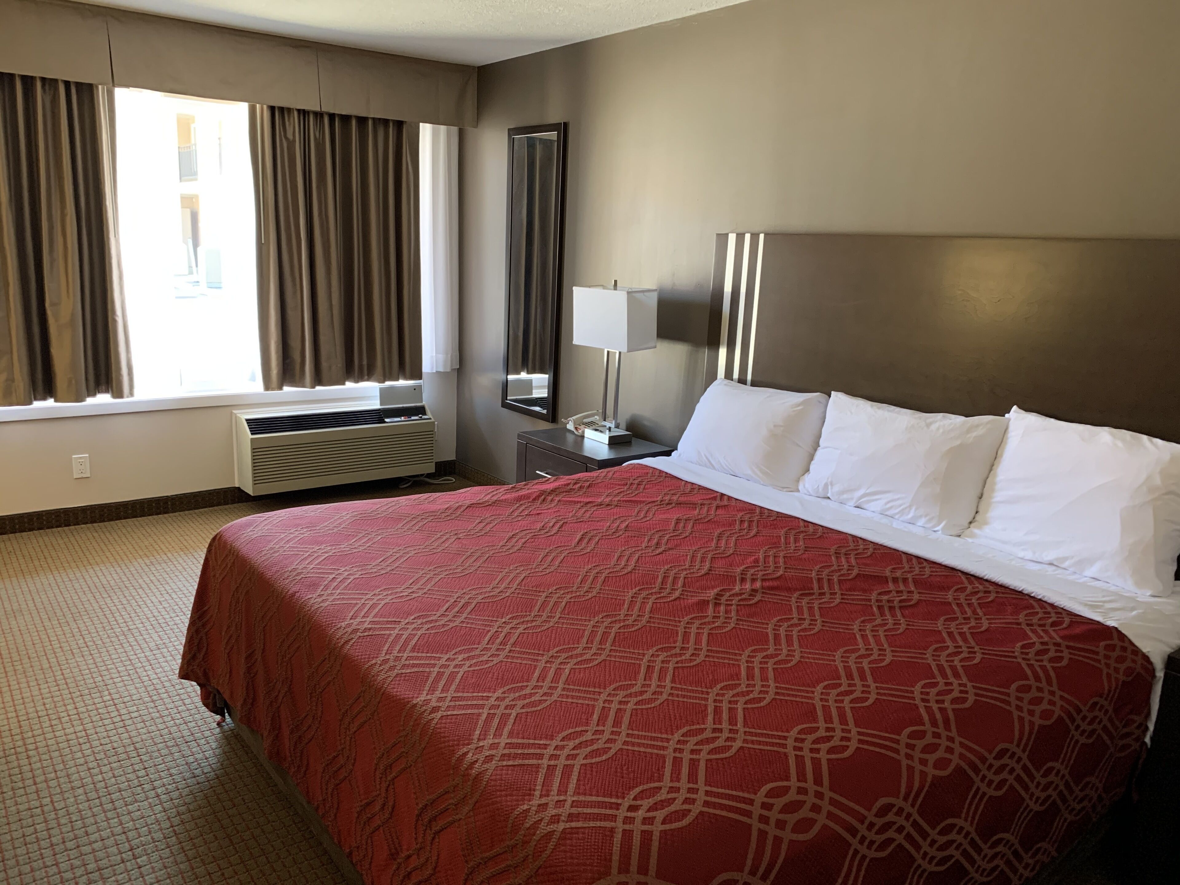 Habitación de hotel con una cama grande, una ventana con cortinas, una mesita de noche con lámpara y un espejo instalado en la pared.