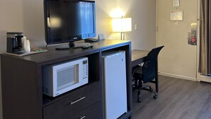A hotel room with a desk, a television, a microwave, a refrigerator, a chair, a lamp, and a door with a lock.