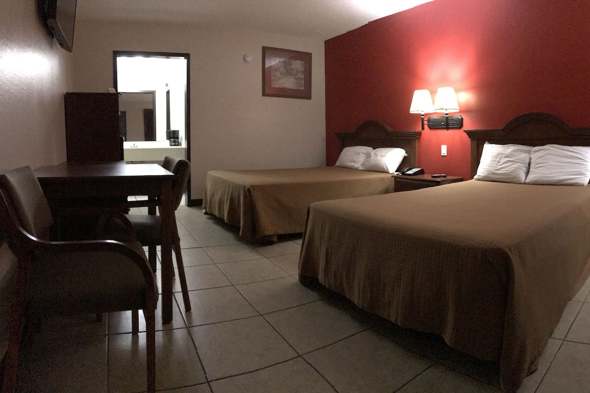 Quarto de hotel com duas camas, uma escrivaninha, cadeiras, telefone e abajur.