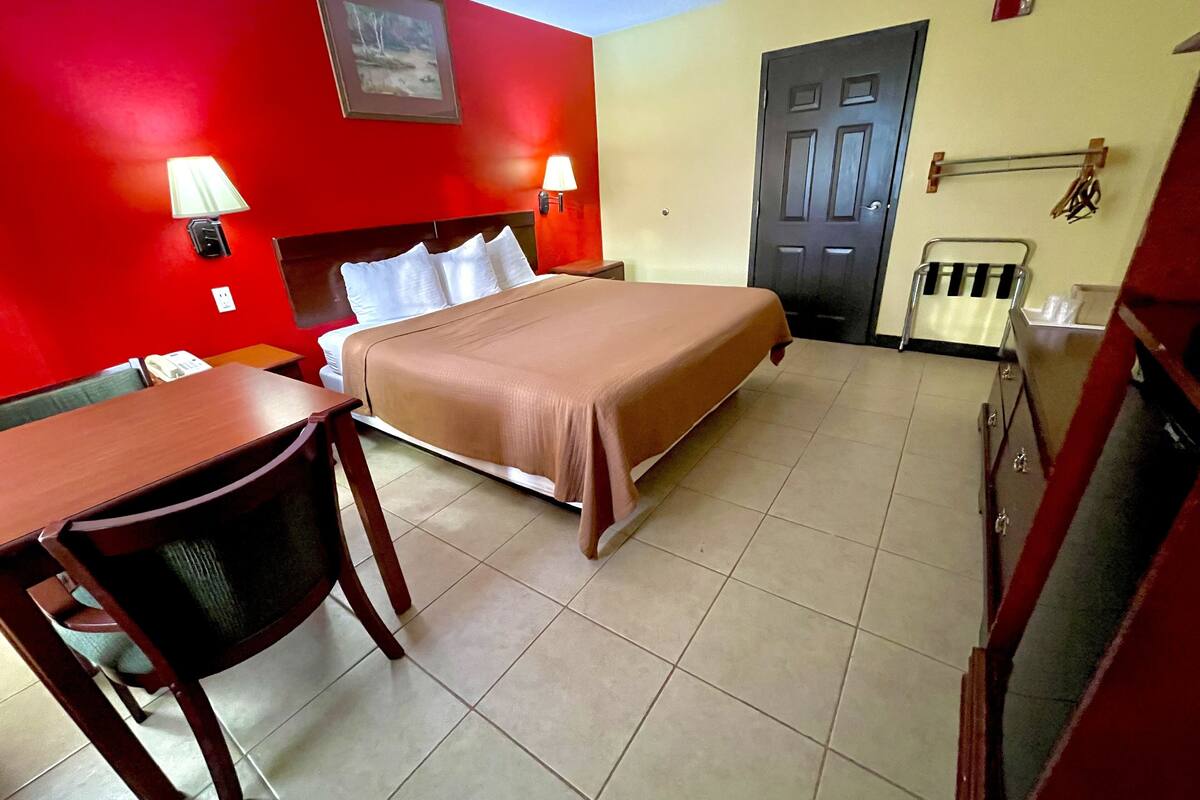Quarto de hotel com cama, escrivaninha, cadeira, luminária, porta e barra de toalhas fixada na parede.