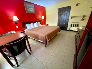 A hotel room with a bed, a desk, a chair, a lamp, a door, and a wall-mounted towel rack.