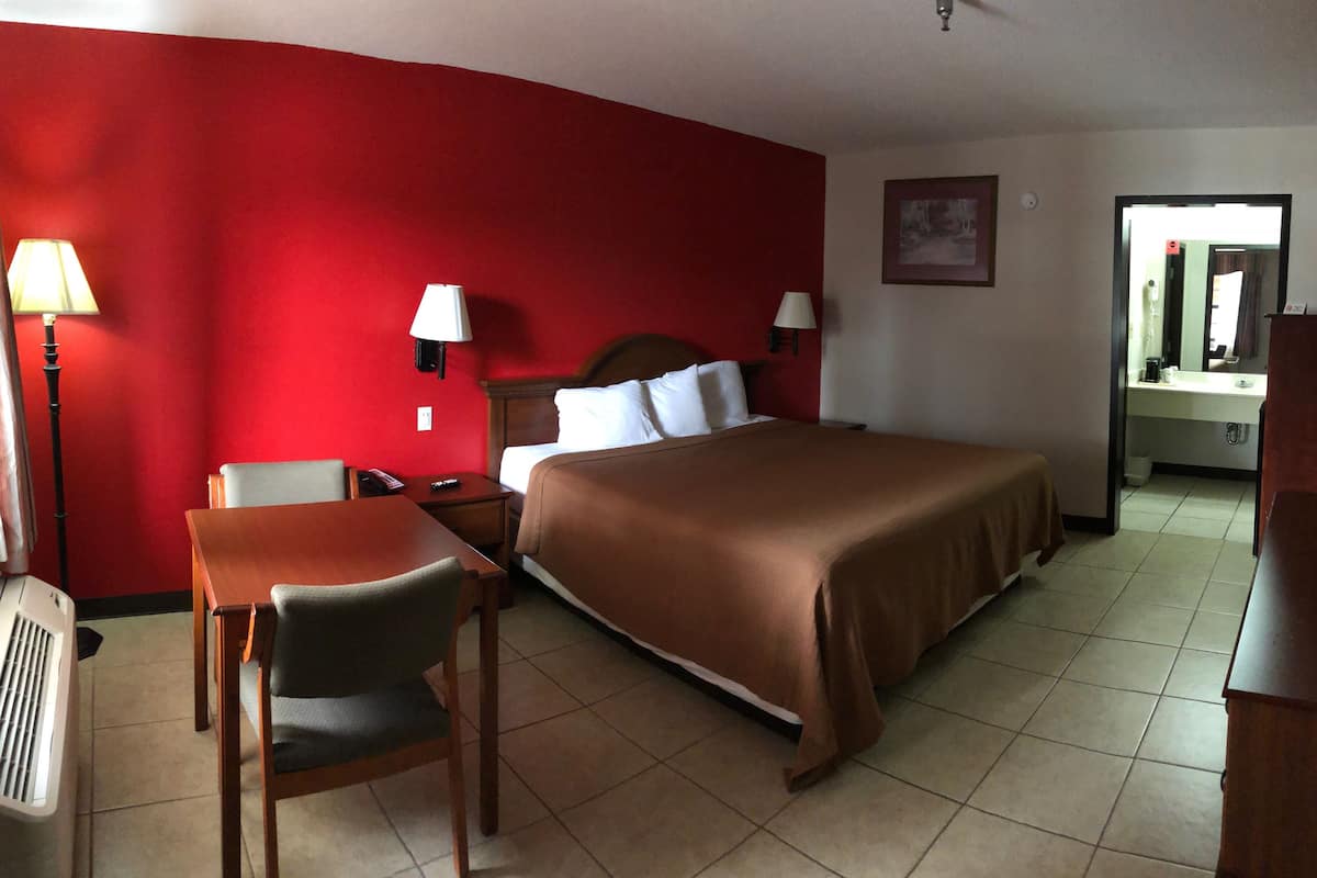Quarto de hotel com parede vermelha, cama com colcha marrom, escrivaninha com cadeira, luminária e espelho.