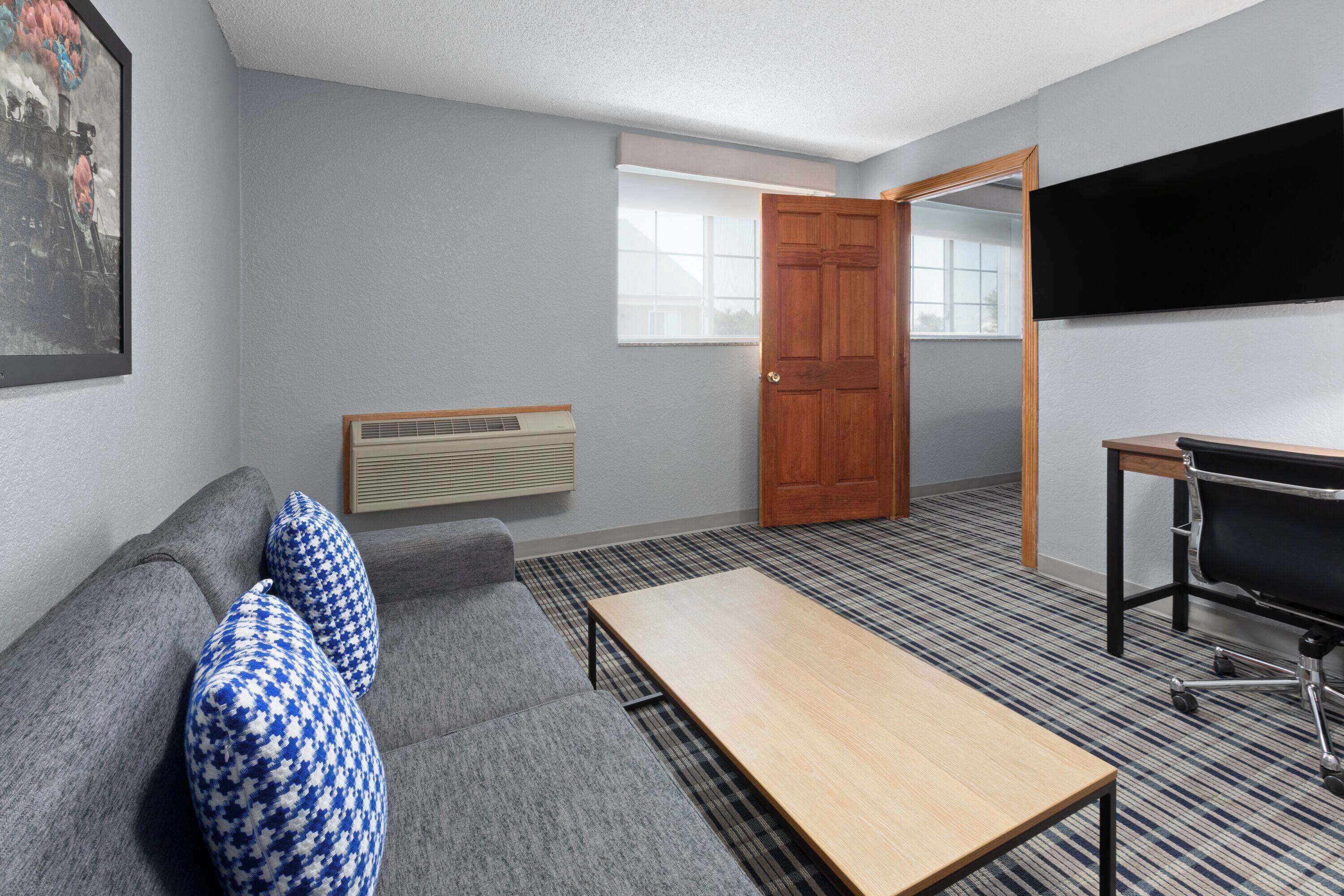 Una habitación con un sofá gris, almohadones estampados, una mesa de madera, una silla de oficina negra, un radiador, una puerta y una ventana.