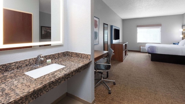 A hotel room with a granite countertop sink, a large mirror, a desk with a chair, a flat-screen TV, a bed, and a window. - AmericInn by Wyndham Grinnell (Grinnell)