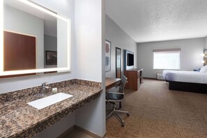 A hotel room with a granite countertop sink, a large mirror, a desk with a chair, a flat-screen TV, a bed, and a window. - AmericInn by Wyndham Grinnell (Grinnell)