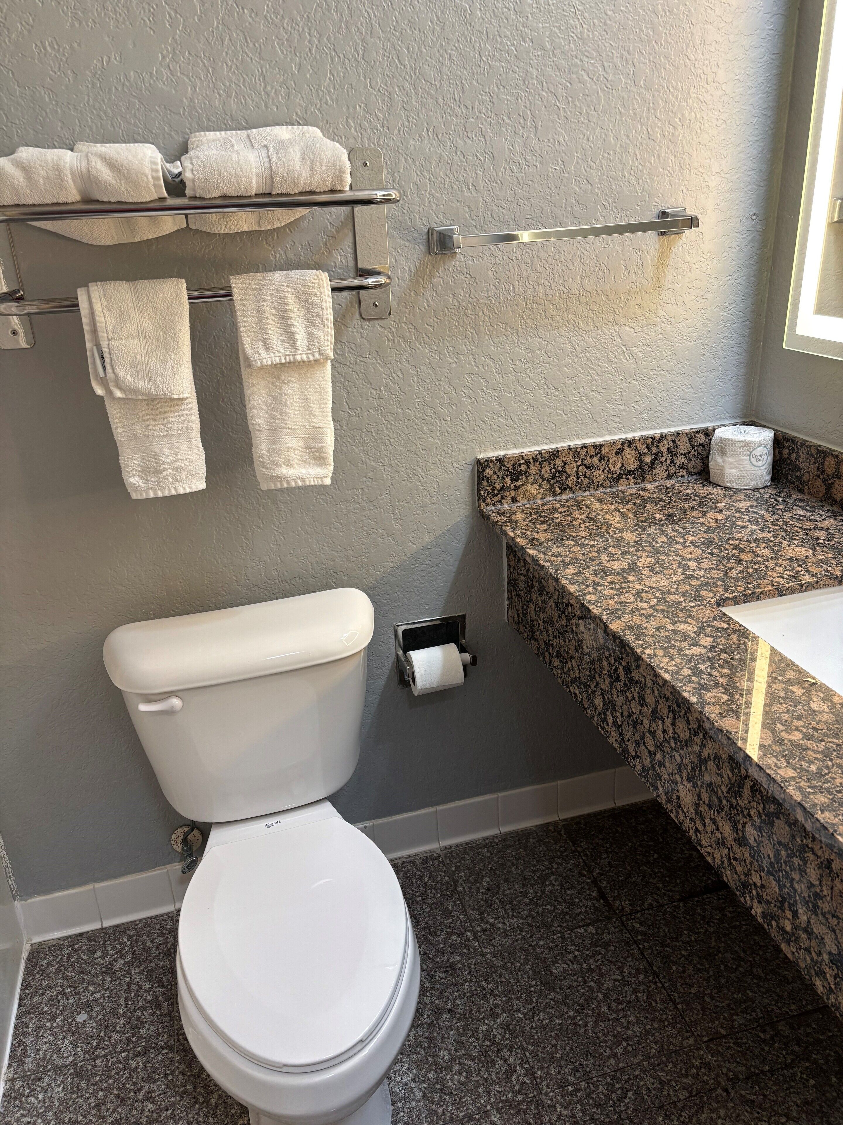 A bathroom with a toilet, a towel rack with towels, a paper towel dispenser, and a granite countertop with a sink.