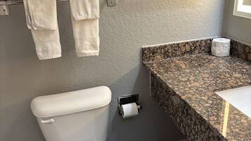 A bathroom with a toilet, a towel rack with towels, a paper towel dispenser, and a granite countertop with a sink.