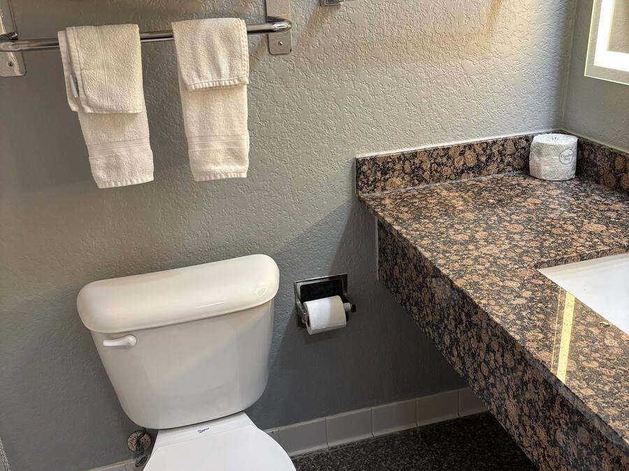 A bathroom with a toilet, a towel rack with towels, a paper towel dispenser, and a granite countertop with a sink.