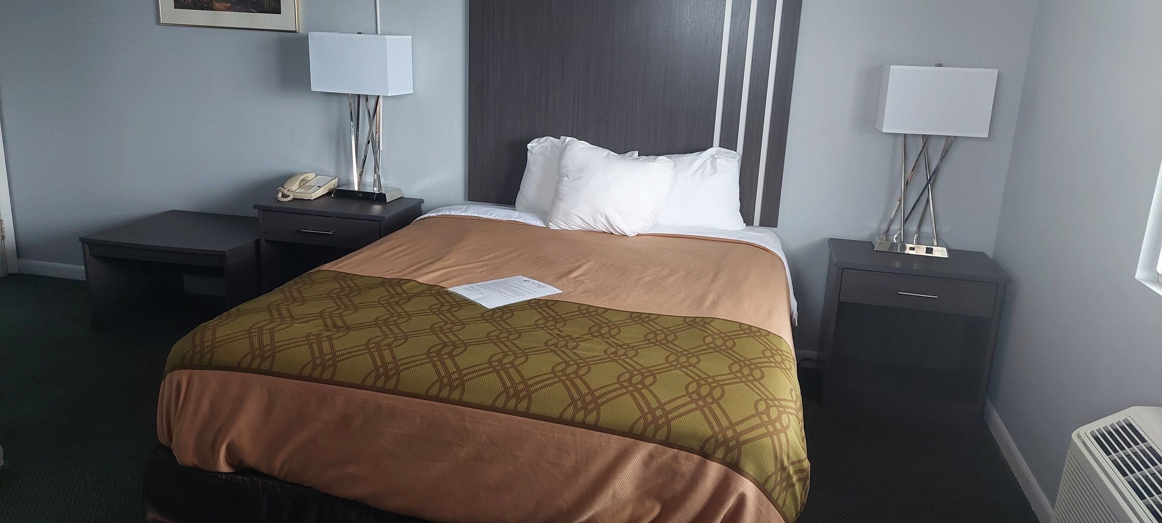 Una habitación de hotel con una cama, dos mesitas de noche, una lámpara, un teléfono y un calefactor.